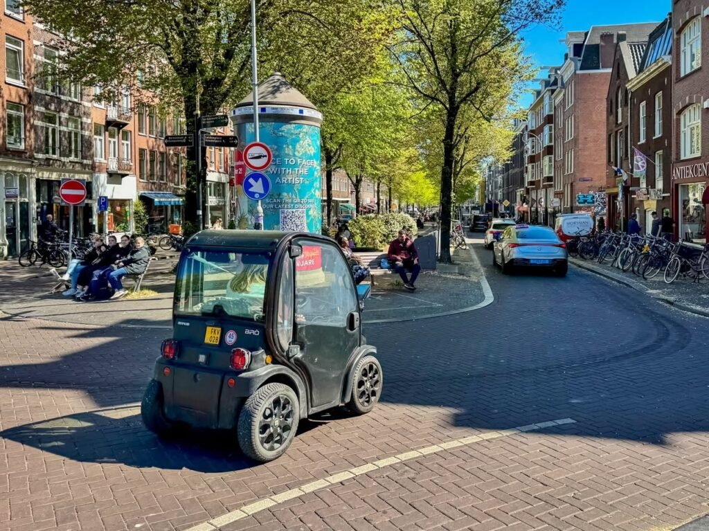A small green electric car on a sunny Dutch street lined with trees and traditional brick buildings.