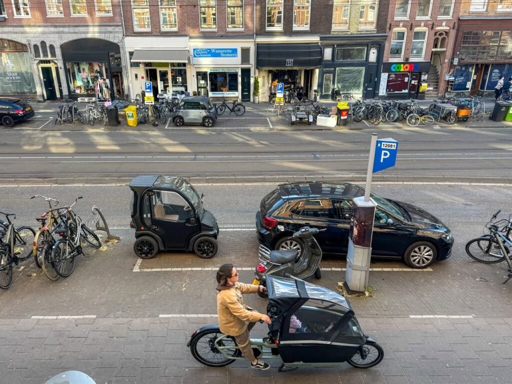 A person rides an e-bike with a child seat through a busy European city street, passing parked bicycles and a black car.