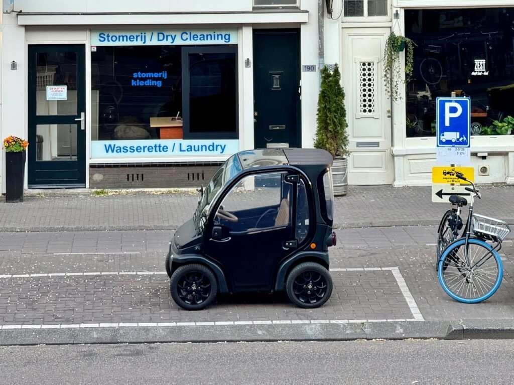 A small black car parked on a cobblestone street next to a dry cleaning shop and bicycle parking sign.