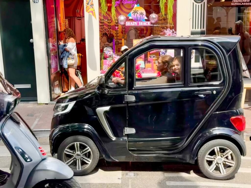 A compact black car parked on a city street near a scooter and storefront window display.