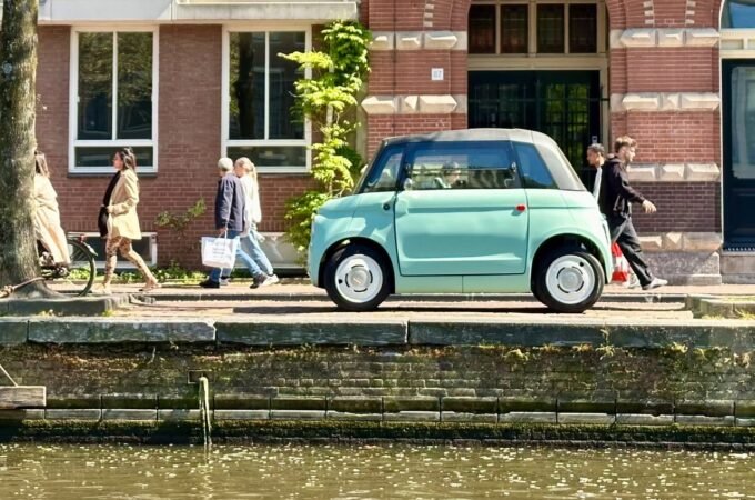 A mint green car parked along a historic canal in Europe. Pedestrians walk past charming brick buildings with red shutters.