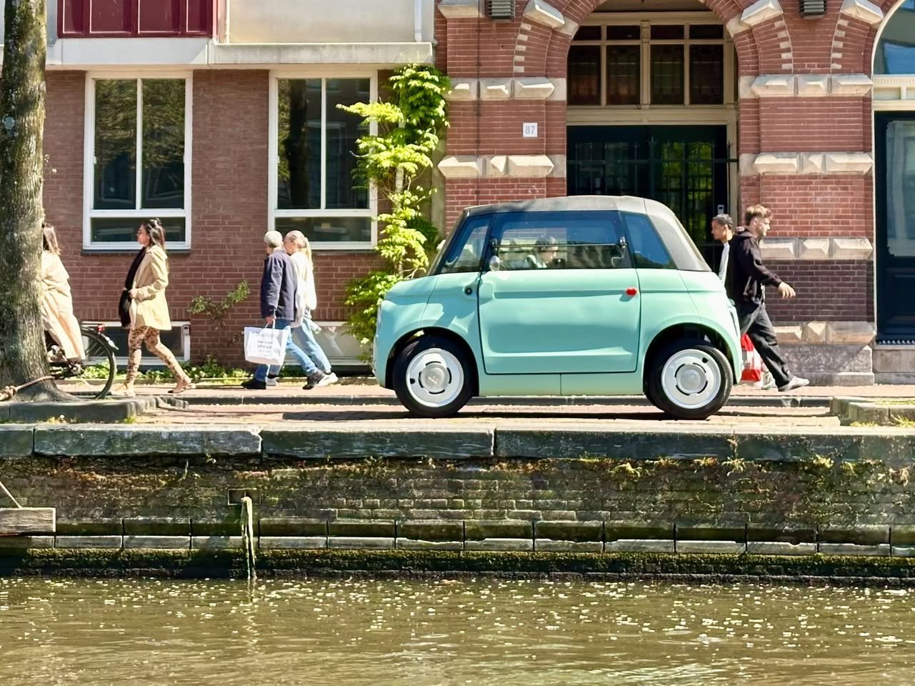 A mint green car parked along a historic canal in Europe. Pedestrians walk past charming brick buildings with red shutters.