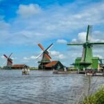Eating Amsterdam Traditional Dutch windmills with their sails catching the wind, set along a calm waterway under a vibrant blue sky.
