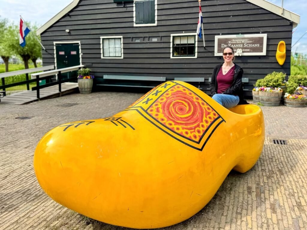 A giant yellow wooden clog sits on a cobblestone patio in front of a dark wooden Dutch building. Dutch flags hanging nearby indicate the location is in the Netherlands.