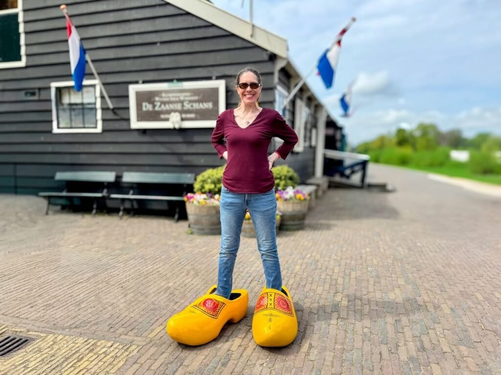 A person stands in giant yellow wooden Dutch clogs outside a building with a Dutch flag.