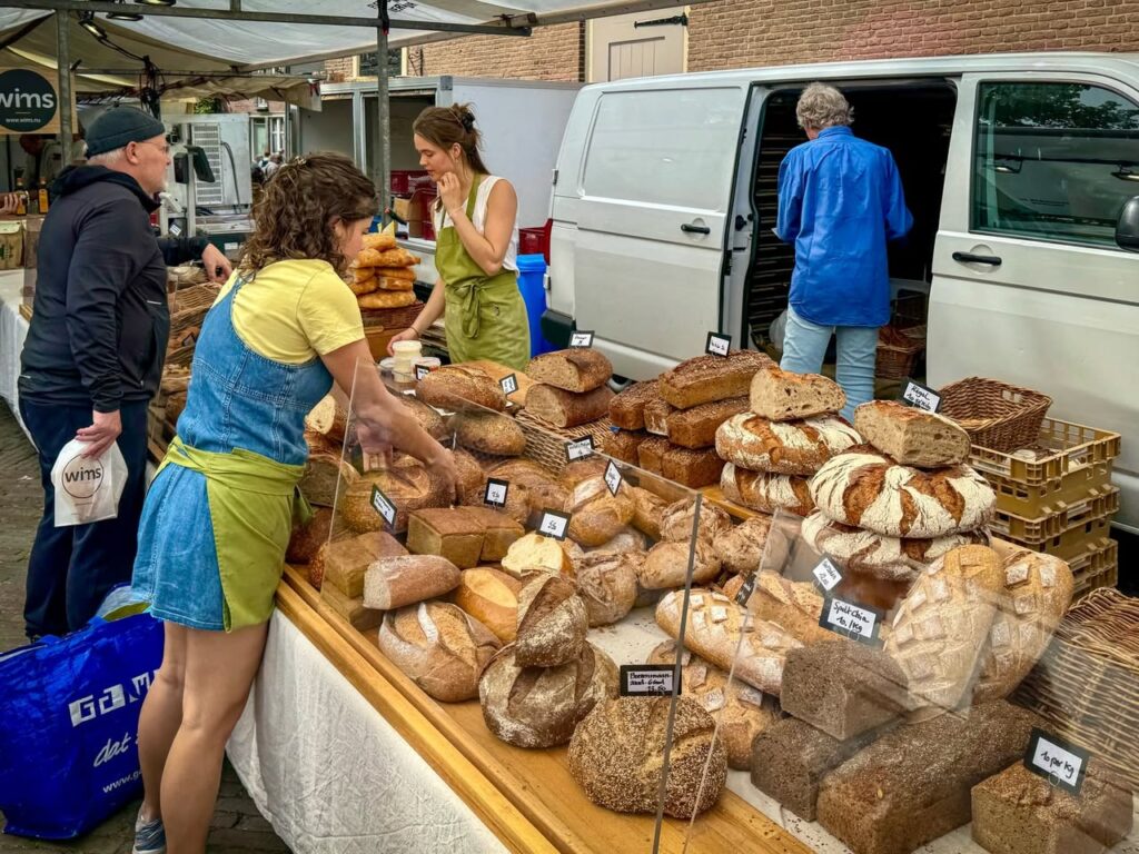 A variety of artisan breads displayed on a table at an open-air market with people shopping nearby.