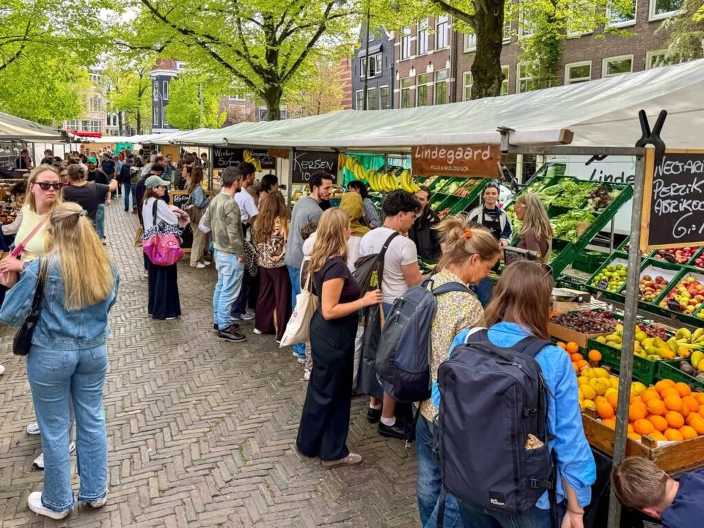 Visitors browse fresh produce at a farmers market in a European town, highlighted by colorful fruits and green trees.