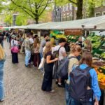 Zaanse Schans Visitors browse fresh produce at a farmers market in a European town, highlighted by colorful fruits and green trees.
