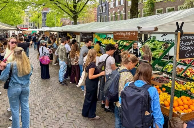Visitors browse fresh produce at a farmers market in a European town, highlighted by colorful fruits and green trees.