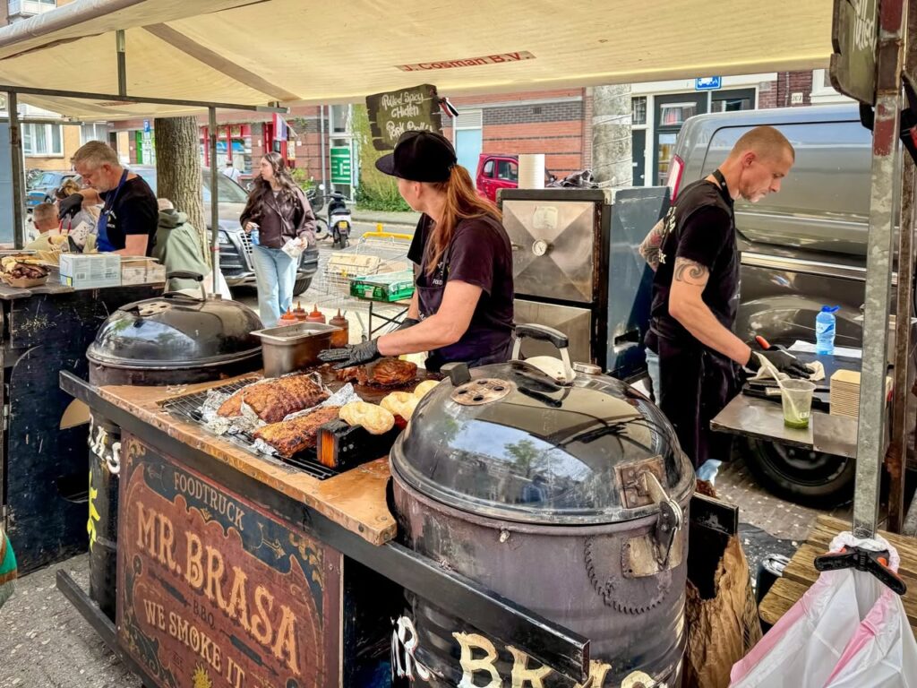 A street food stand named Mr. Brasa preparing grilled meats on large drums at an outdoor market. Vendors in dark shirts work at the cook station while customers browse nearby.
