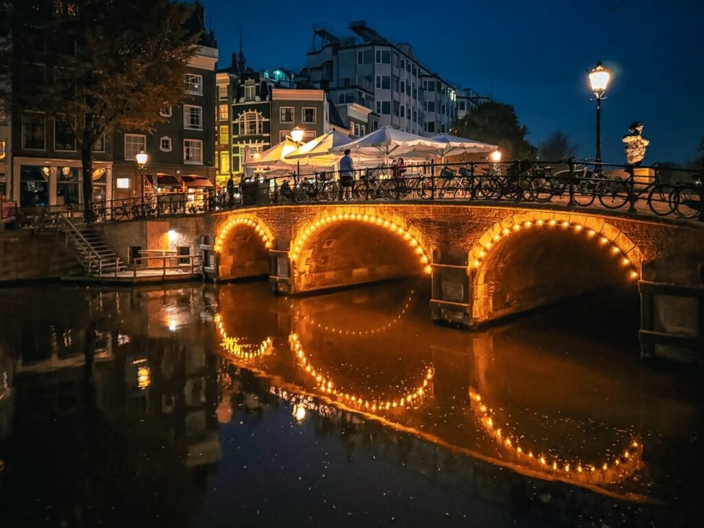 A stunning night view of a historic stone bridge in Amsterdam, with warm lights reflecting beautifully on the canal. String lights adorn the arches, highlighting the bridge's elegant design against the dark water.