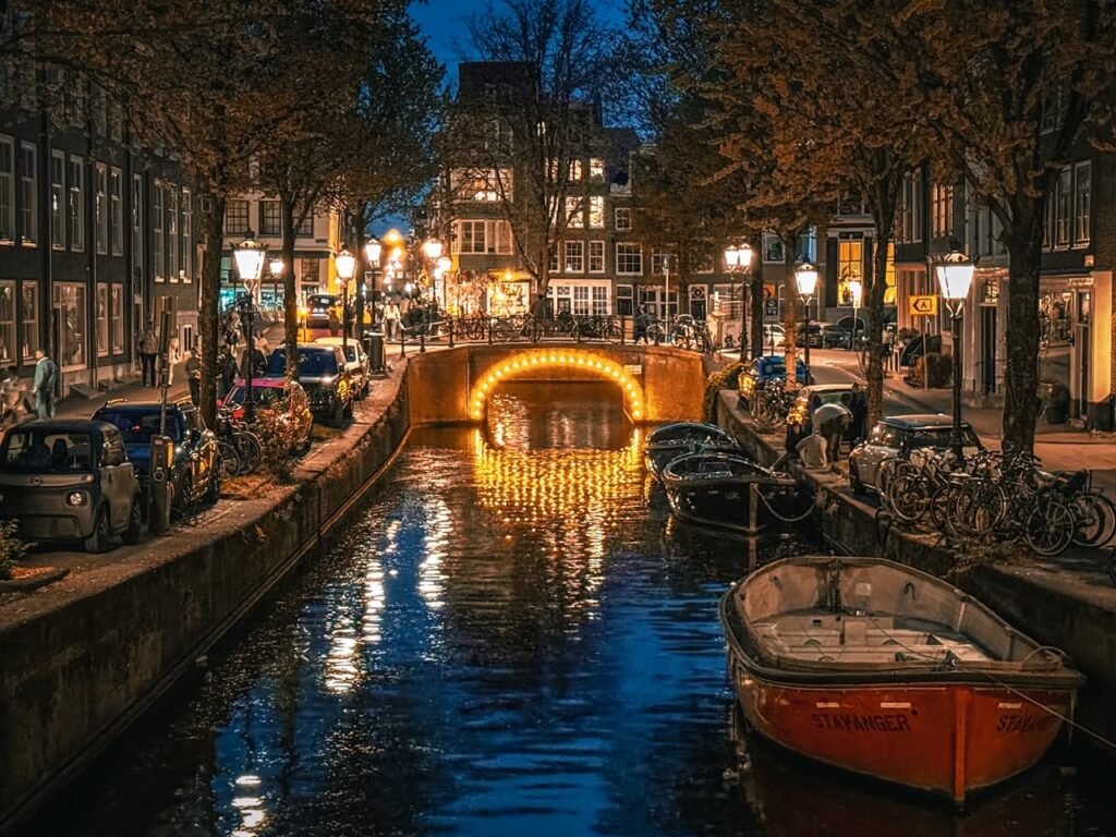 A serene twilight scene featuring a canal lined with traditional Dutch buildings and boats. A warm-lit bridge illuminates the water with glowing reflections.