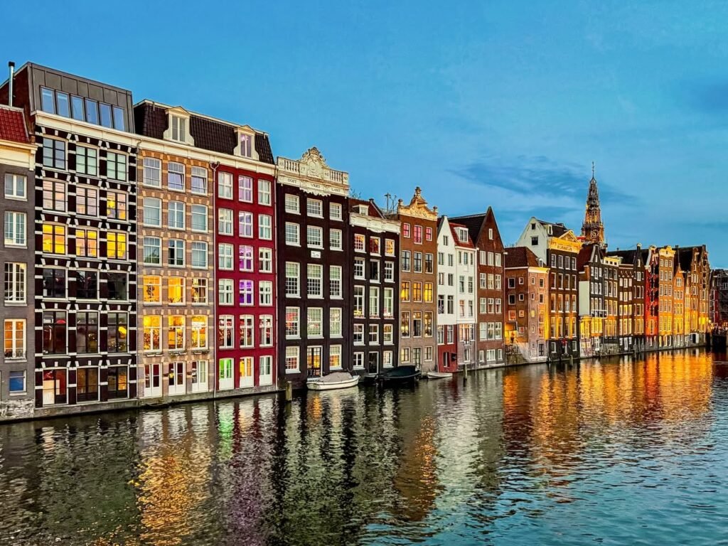 Colorful traditional Dutch houses line an Amsterdam canal with warm reflections on the water. A church spire is visible in the blue evening sky.