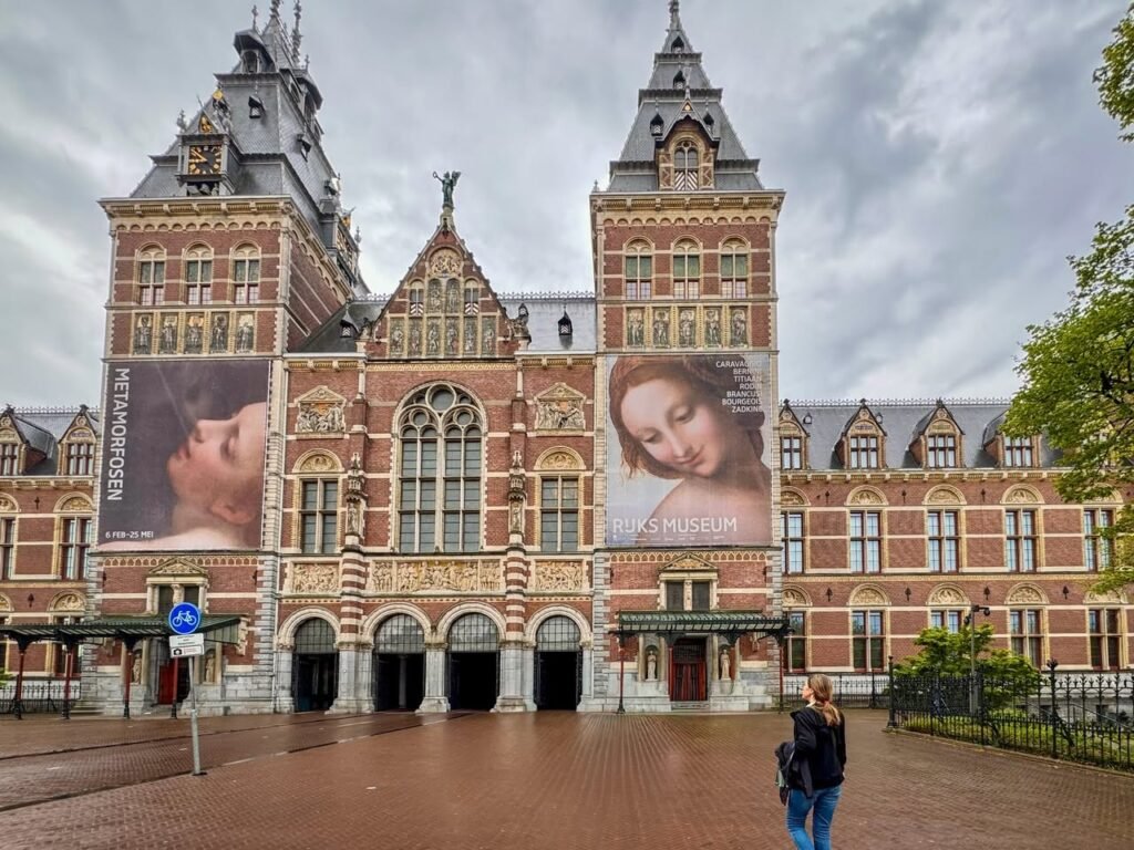 The exterior of the Rijksmuseum in Amsterdam, featuring its iconic red-brick facade and distinctive towers under a cloudy sky.