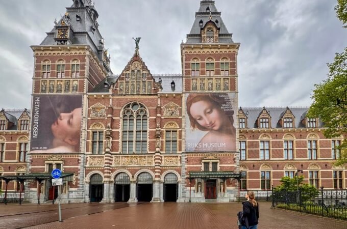 The exterior of the Rijksmuseum in Amsterdam, featuring its iconic red-brick facade and distinctive towers under a cloudy sky.