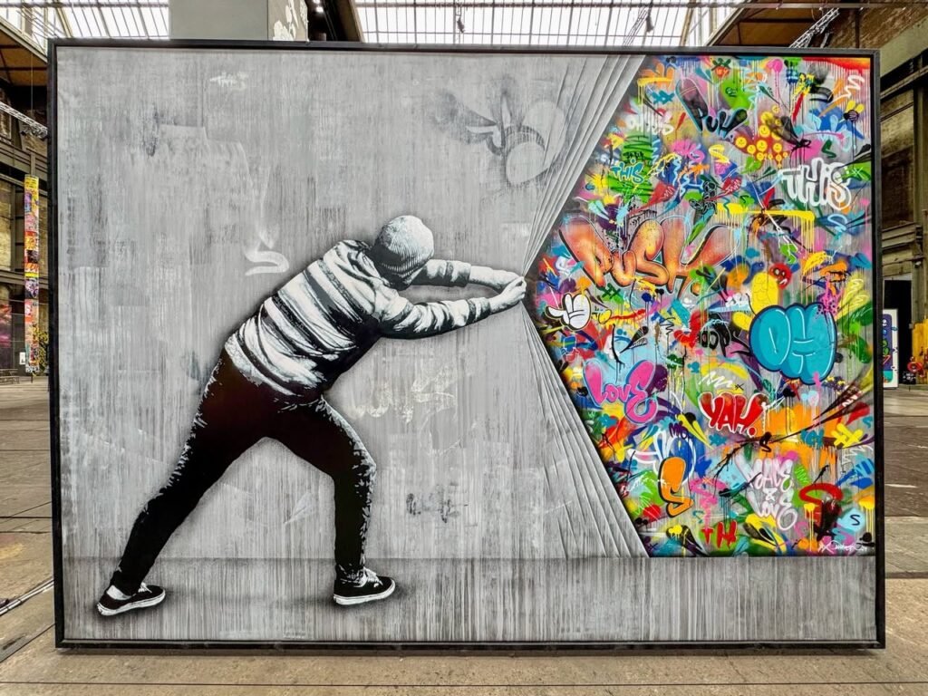 A figure pushes an easel to reveal a colorful abstract design. This large-format artwork blends monochrome and vibrant hues in an industrial setting.
