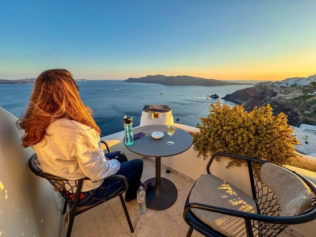 A person enjoying a drink on a tranquil balcony overlooking the Aegean Sea in Santorini, Greece. The warm sunset colors highlight the coastal landscape and scenic beauty.
