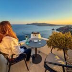 STRAAT Museum A person enjoying a drink on a tranquil balcony overlooking the Aegean Sea in Santorini, Greece. The warm sunset colors highlight the coastal landscape and scenic beauty.