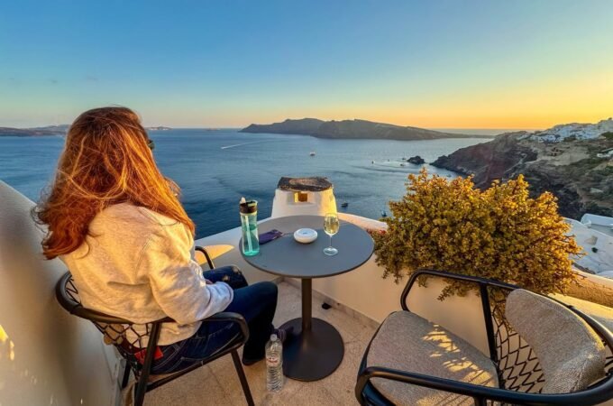 A person enjoying a drink on a tranquil balcony overlooking the Aegean Sea in Santorini, Greece. The warm sunset colors highlight the coastal landscape and scenic beauty.