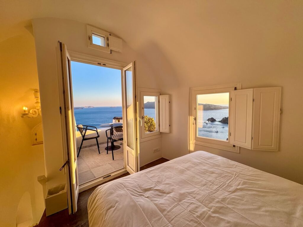 A cozy bedroom featuring an open balcony door and windows overlooking the ocean. The room is bright with natural light and views of the water.