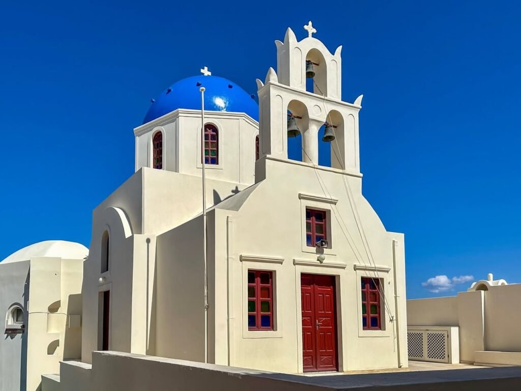 A traditional white Greek church with blue domes and a red door sits against a vivid blue sky. The close-up details highlight the bell tower and architectural design.