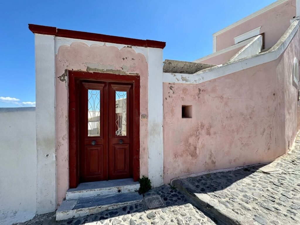 A warm pink building with a rich red wooden double door on a sunny cobblestone street.