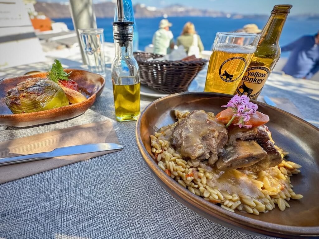 Grilled bread with olives and a plate of meat on orzo served in a rustic wooden bowl, alongside a glass of Yellow Tail beer on a table by the water.