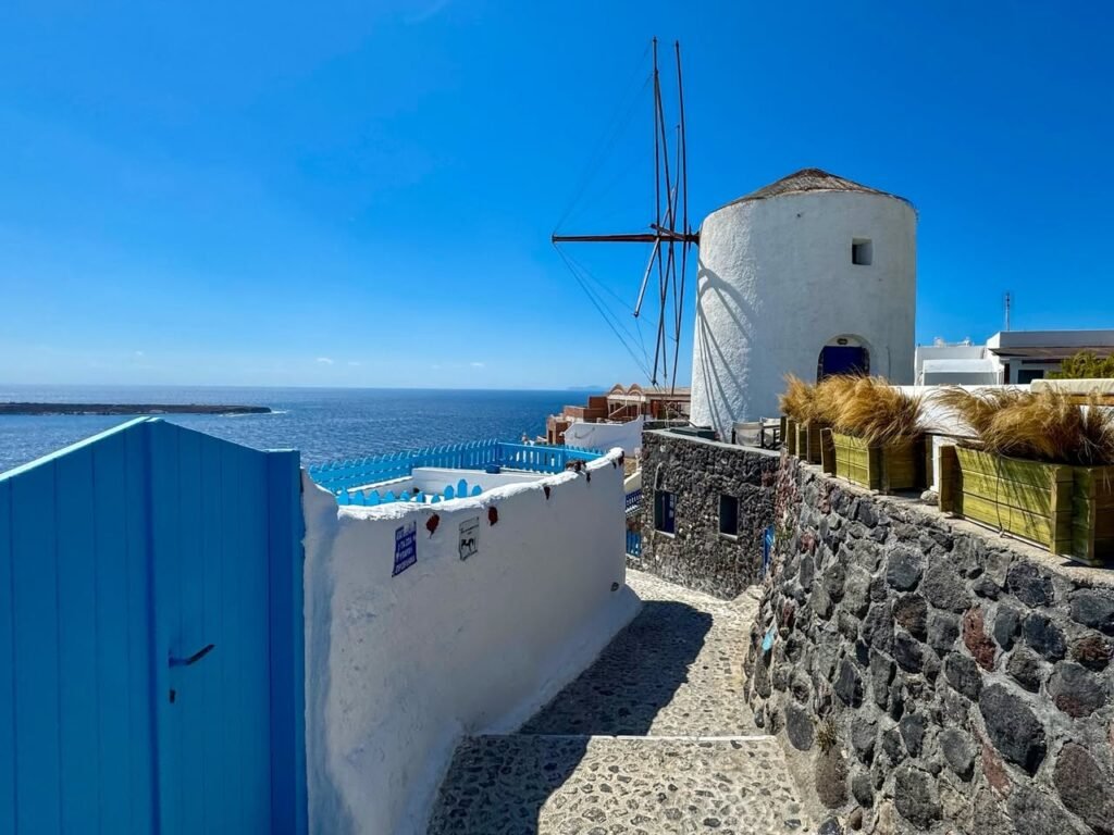 A picturesque scene featuring a traditional white windmill and blue walls in Santorini, overlooking the deep blue sea under a clear sky.