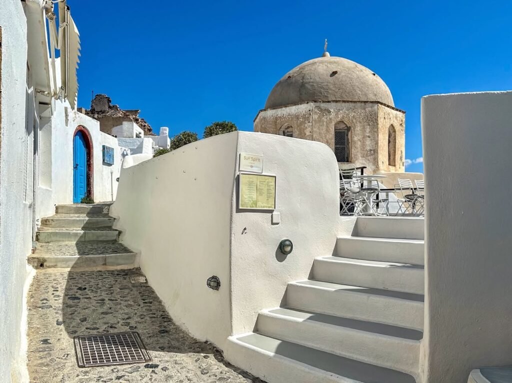Scenic view of whitewashed walls, stone steps, and a historic domed church. A cobblestone path leads through a sunlit courtyard under clear blue skies.