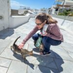A traveler crouches down to interact with a small, stray cat on a bright, sunny terrace with coastal architecture visible in the background.