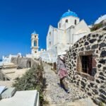 Cats of Santorini A person walks a stone path beside historic dark stone walls leading to a white church with a blue dome in the Greek islands.