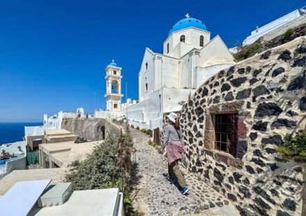 A person walks a stone path beside historic dark stone walls leading to a white church with a blue dome in the Greek islands.