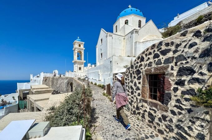 A person walks a stone path beside historic dark stone walls leading to a white church with a blue dome in the Greek islands.