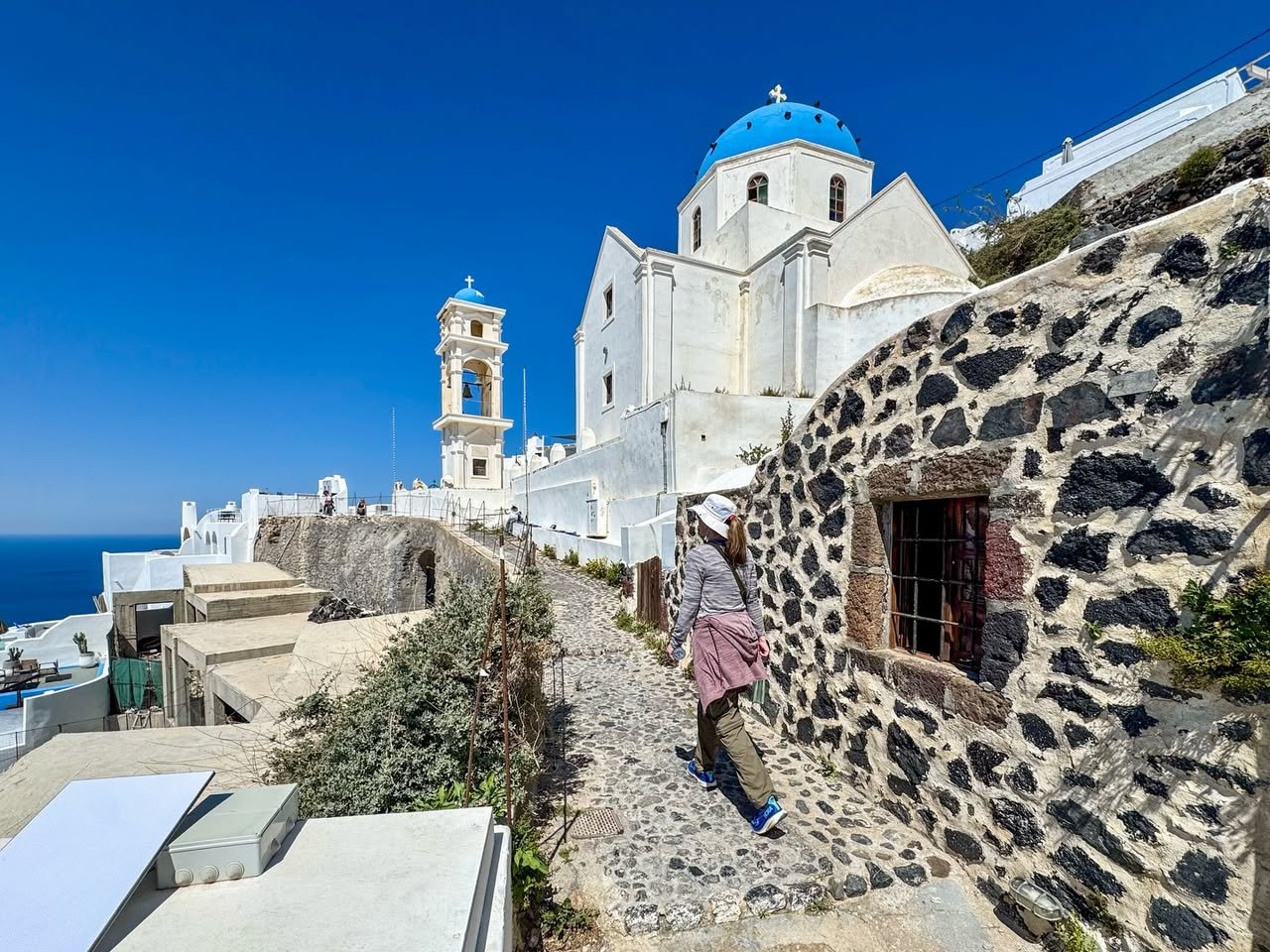 Fira to Oia Caldera Hike A person walks a stone path beside historic dark stone walls leading to a white church with a blue dome in the Greek islands.