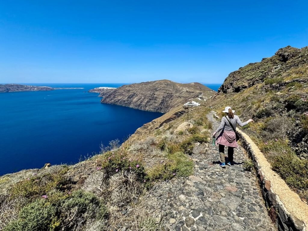 Hiker walking along a rugged stone castle walkway overlooking deep blue ocean water under a clear blue sky.