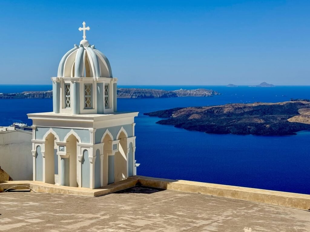 A small white domed chapel with a cross overlooks the deep blue sea and distant islands.