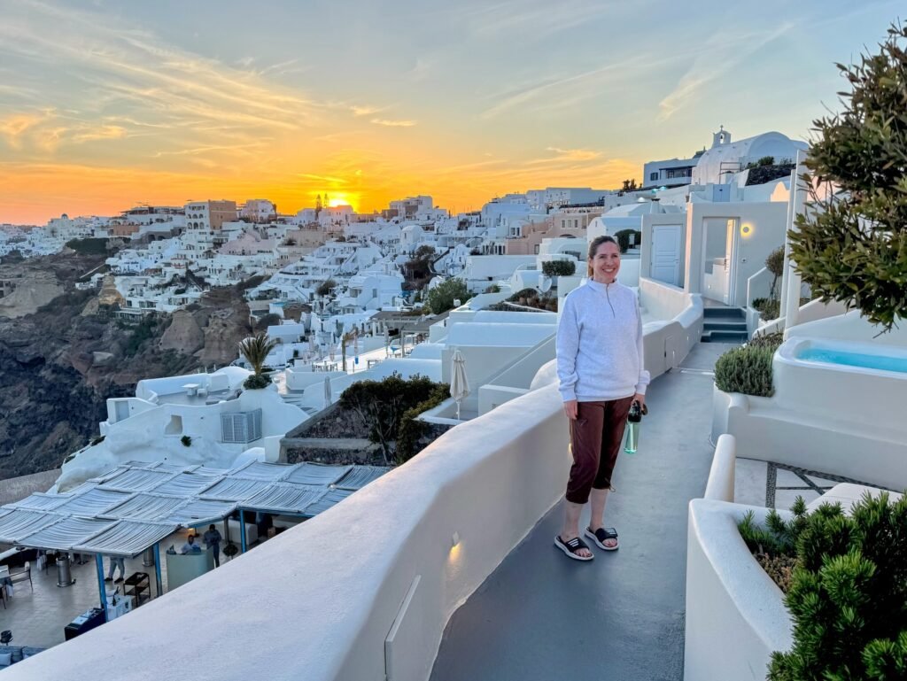 A traveler relaxes on a terrace overlooking white-washed buildings in Santorini, Greece, against a golden sunset sky.