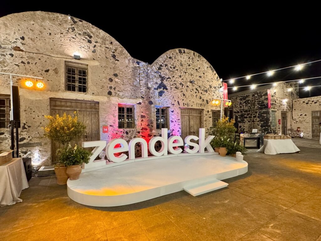 A nighttime view of a large Zendesk sign and raised platform in front of a unique stone building with arched windows. String lights decorate the courtyard.