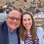 Athens A selfie captured at a lively outdoor event with a rustic stone wall backdrop. Guests are seated at tables enjoying the atmosphere.
