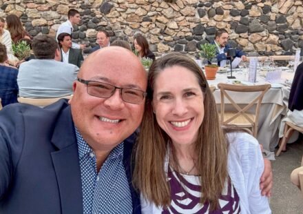 A selfie captured at a lively outdoor event with a rustic stone wall backdrop. Guests are seated at tables enjoying the atmosphere.