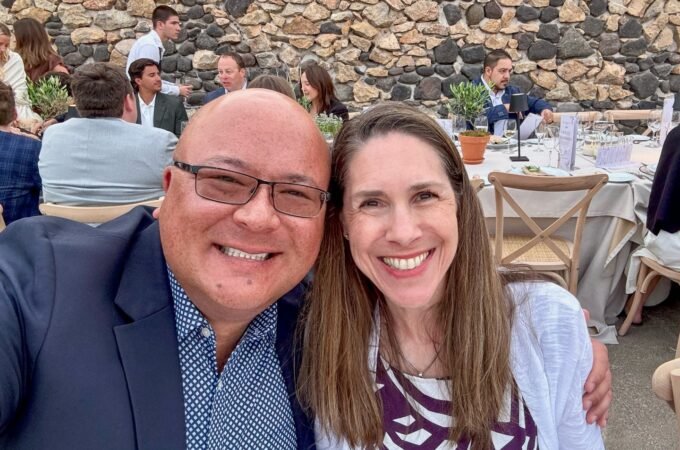 A selfie captured at a lively outdoor event with a rustic stone wall backdrop. Guests are seated at tables enjoying the atmosphere.