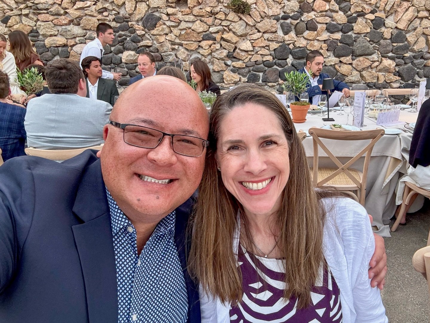 A selfie captured at a lively outdoor event with a rustic stone wall backdrop. Guests are seated at tables enjoying the atmosphere.