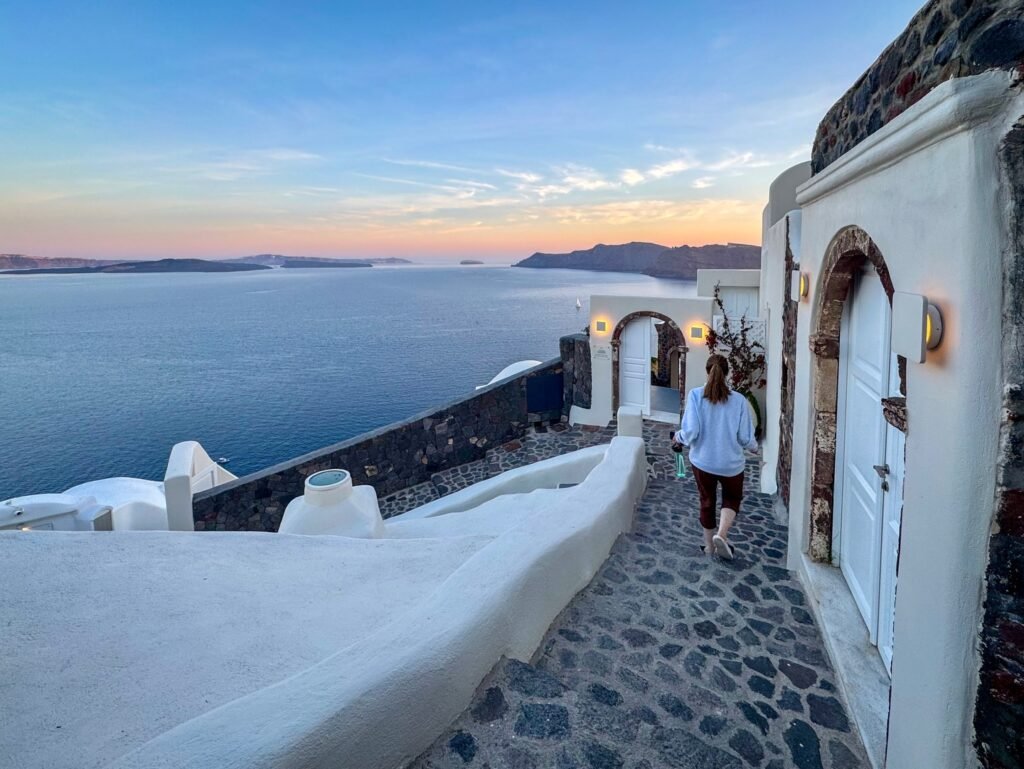 A pebbled pathway winds through white-washed Cycladic architecture with a stunning Aegean Sea view at twilight.