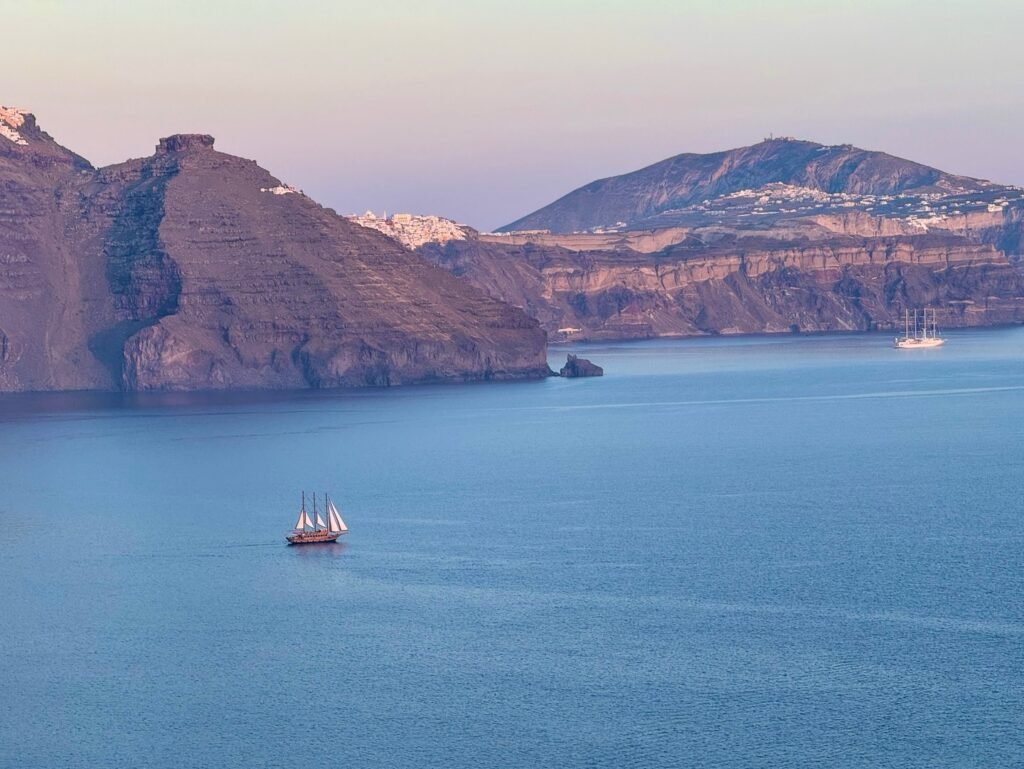 A traditional sailing ship on the blue Aegean Sea. The cliffs of Santorini rise in the background under a pastel sky.