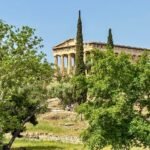 Ancient Greek temple atop a hill surrounded by lush green trees and cypress, under a clear sky.