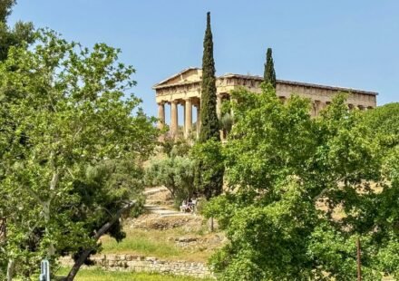 Ancient Greek temple atop a hill surrounded by lush green trees and cypress, under a clear sky.