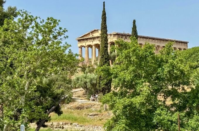 Ancient Greek temple atop a hill surrounded by lush green trees and cypress, under a clear sky.