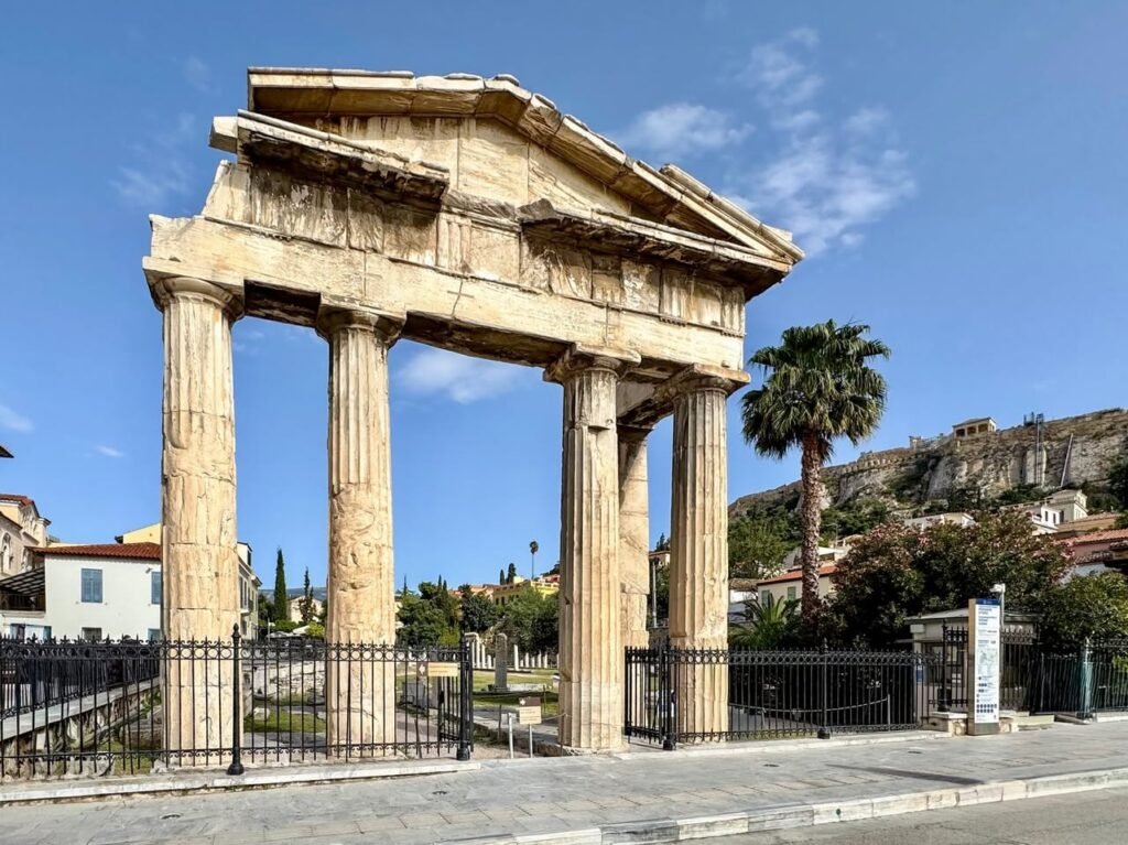 A Greek temple structure featuring large columns and a pediment, set against a clear blue sky under a bright sun. The stone facade and architectural details are showcased in the daylight, surrounded by a metal fence and clear sky.