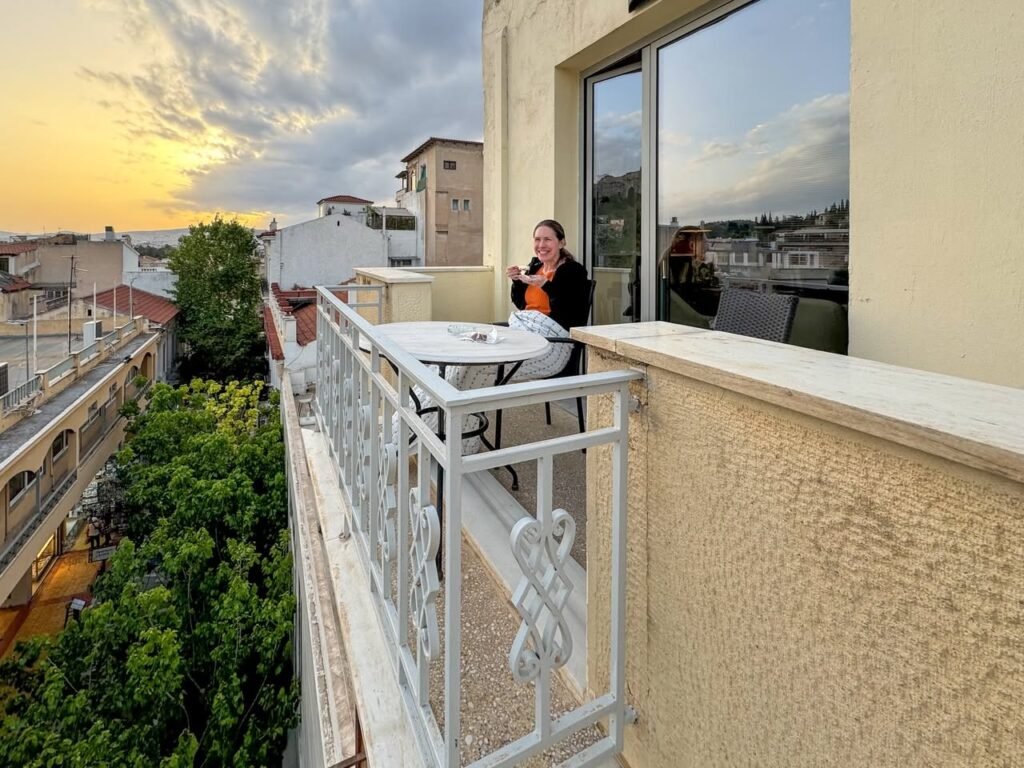 A person stands on a balcony with a table, overlooking historic architecture at sunset.