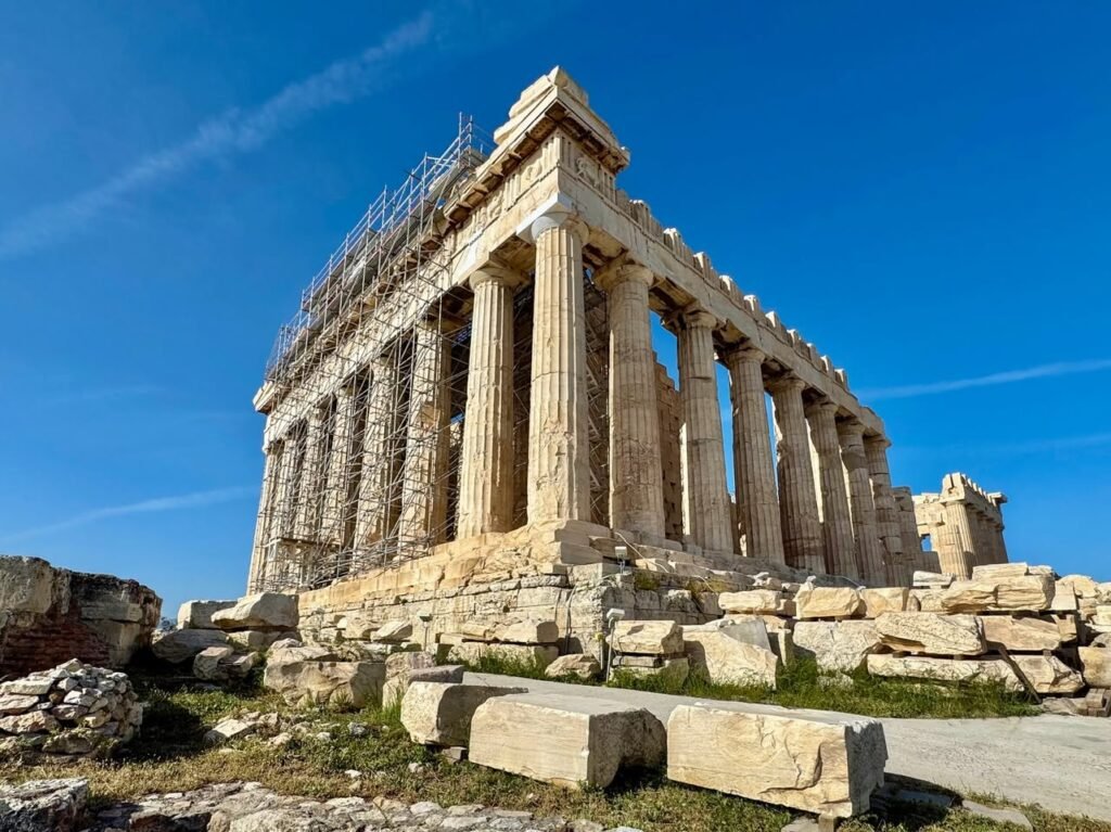 A classic Greek temple with detailed columns stands against a vivid blue sky. Scattered ruins and stone blocks add to the historic ambiance.